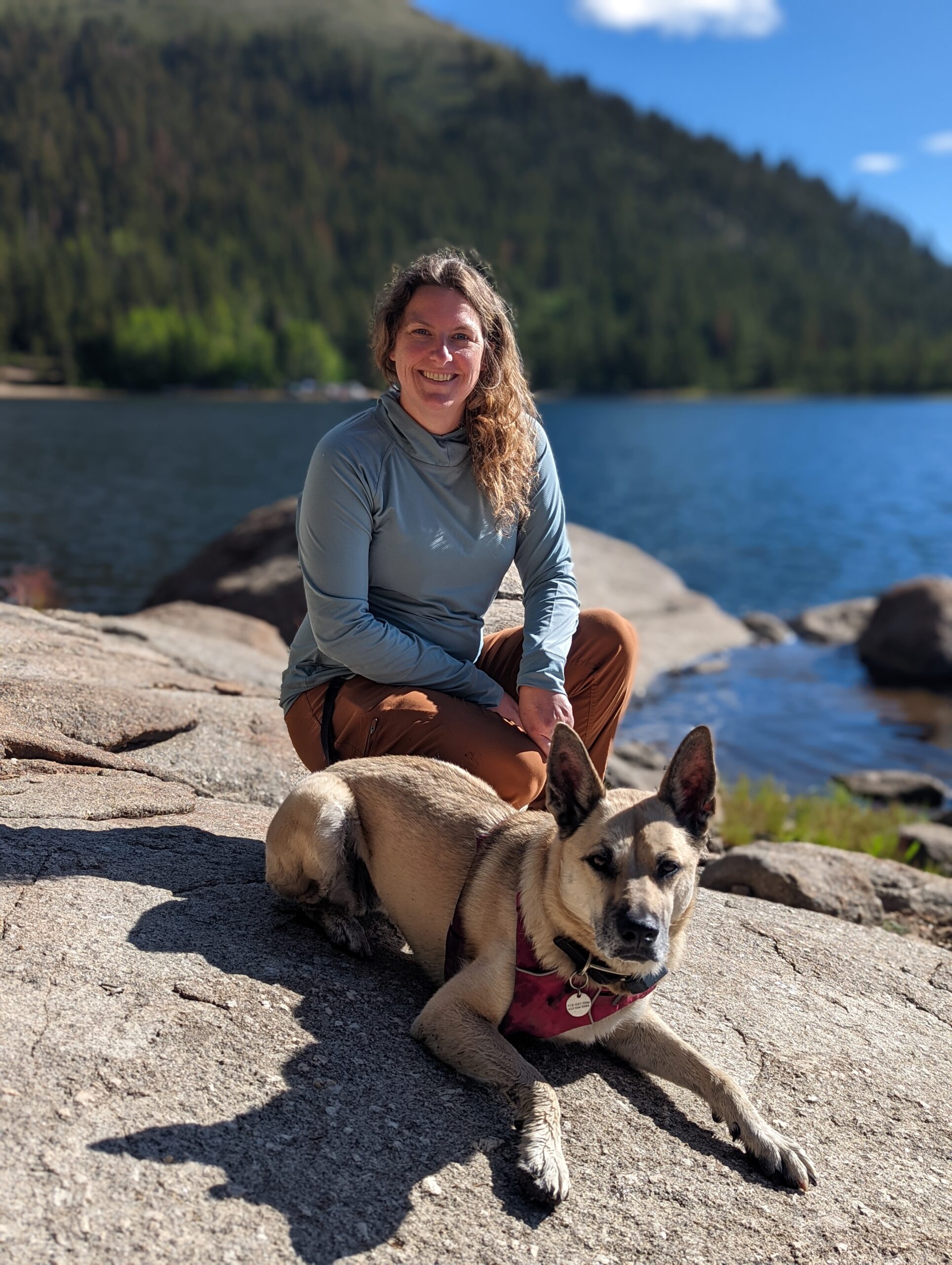 Beth sits on a granite slab of stone with her german shepherd mix. A lake and mountain are in the background.